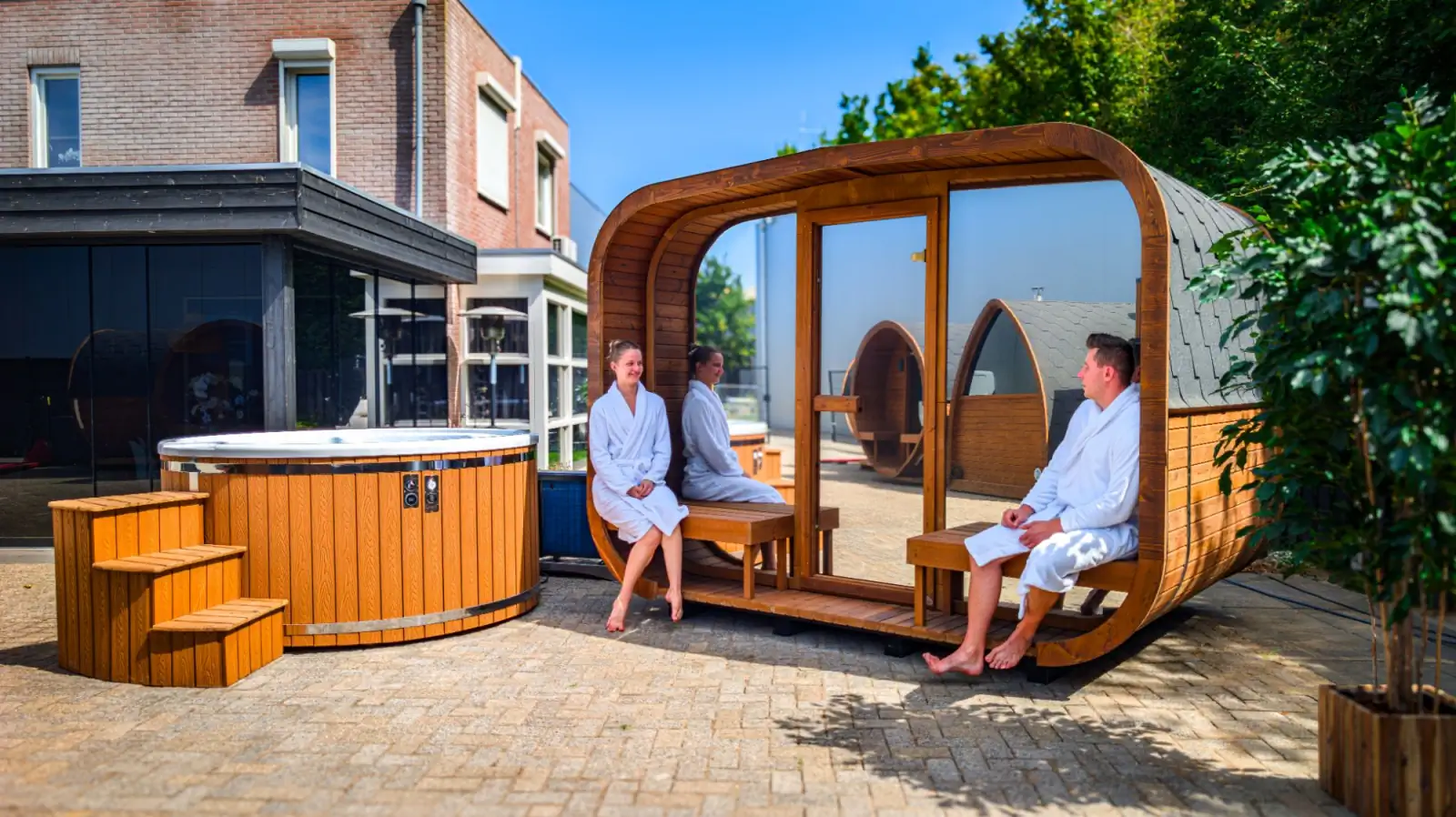 Is een hybride hottub de meerprijs waard? | Hottub en buitensauna Hottub met houten trap naast een buitensauna met glazen voorkant, mensen in badjas op het terras