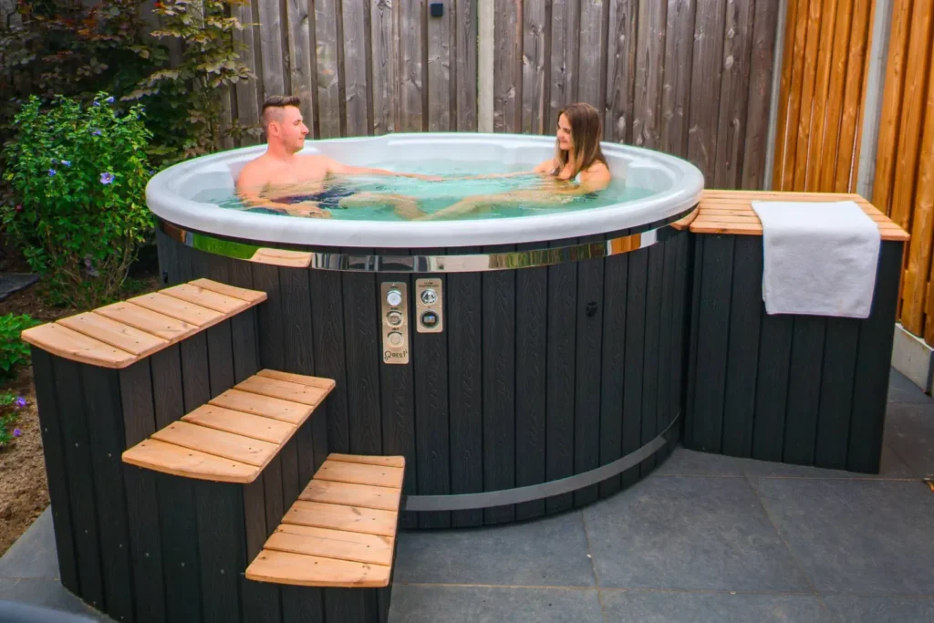 Un hot tub hybride vaut-il le supplément de prix ? | Hot tub dans le jardin Deux personnes dans un hot tub rond avec bordure noire et escalier en bois dans un jardin
